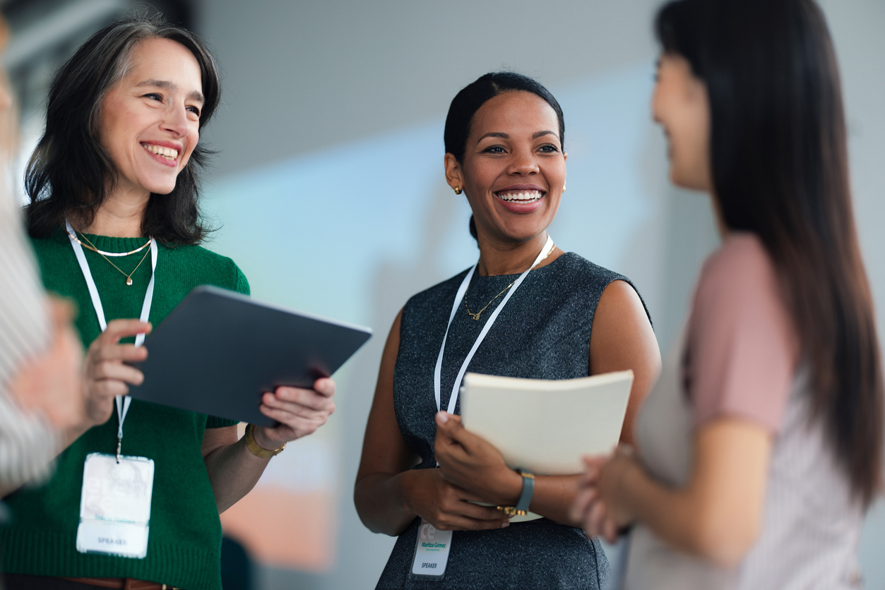 A group of smiling professionals mingle at a conference, sharing ideas and materials. Lanyards and badges indicate a formal event, emphasizing collaboration, learning, and networking.